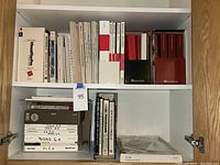 Wide view of cabinet shelves with stacked vintage technical manuals, mostly white and red spined books, including Microsoft and AutoCAD manuals.