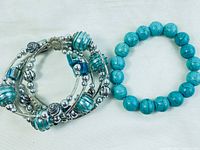 Two bracelets shown: one is a blue stone bead bracelet, the other a multi-strand bracelet with turquoise and silver-toned glass beads and metallic accents, lying on white fabric.
