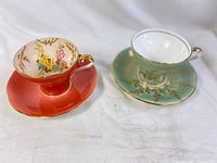 Two vintage Aynsley teacup sets shown from side angle on white background, one coral with floral design and one turquoise with gold detailing.