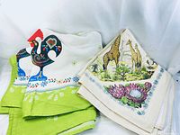 Both tablecloths folded and displayed showing main decorative motifs: colorful Portuguese rooster and African giraffes with flowers.