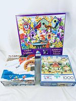 Three puzzle boxes displayed on a light surface: Garden Neighbors, Charles Wysocki winter village, and carousel puzzle.