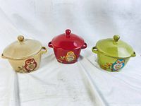 Front view of three owl-themed ceramic soup dishes in beige, red, and green with lids on a white background.