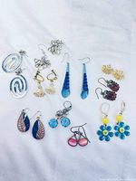 Full view of all 13 pairs of earrings showing varied styles, colors and materials on white fabric background