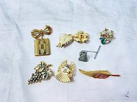 Eight vintage brooches arranged on white cloth showing various gold-tone and silver-tone pins with decorative details including a bow, flower, grapes, heart, fan, iron, feather, and rectangular pendant.