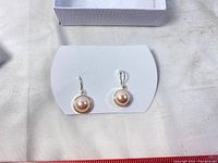Pair of pale pink faux pearl button drop earrings on white card, showing front view and details of pearl and metal bezel.