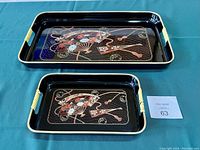 Two black Japanese lacquer trays with floral fan designs placed side by side to show size difference and detail.