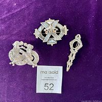 Set of three Celtic or Nordic style brooches arranged on purple fabric background, including one oval cross brooch with stones, one bird motif brooch, and one slender twisted pin.