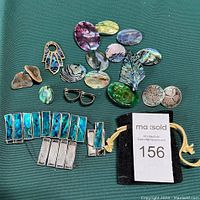 Overview of the lot showing abalone shell pieces for earrings, pins, and a silver-toned bracelet with blue abalone inlays along with a black pouch showing lot number.