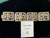 Top view showing 4 decorative linked panels of brass cuff bracelet with fleur de lis, crowned shield, knight on horseback, Cupid cherub motifs.