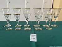 Overview of 11 stemware pieces, 6 large goblets and 5 smaller glasses, arranged on green cloth showing gold rims and stems.