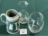 Three items displayed on green cloth: a Dudson green jasperware jug with white classical relief details, a clear glass vase with angular faceted rim, and a clear round glass vase.