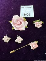 Overall view showing all four jewelry pieces on a purple velvet background: a large rose brooch, matching rose stud earrings, a small stud pin, and a stick pin with rose and leaves.