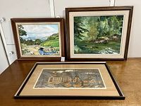 Three framed artworks on a wooden table, showing two landscape paintings and one woodcut print.