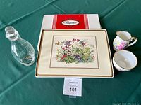 Clear image showing glass cruet with lid, two-piece cream and sugar set with floral pattern and gold trim, and one of the Pimpernel placemats with red and white Pimpernel box behind.