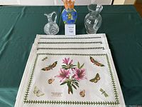 Four cloth Portmeirion placemats displayed stacked with a cut glass cruet, colorful floral vase, and clear glass vase behind them on a green tablecloth.