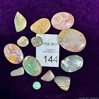 Group of 13 polished stones on a purple background, various colors and sizes shown with a lot number card.