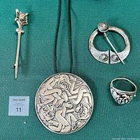 Overview of all four jewelry pieces arranged on green fabric: vintage rose ring, round pendant on leather cord, two silver cloak pins including IONA pin.
