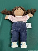 Front view of the doll with brown yarn pigtails, wearing pink and white striped shirt, blue jeans, and white shoes with blue detailing.