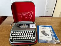 Front view of Smith Corona Skywriter portable typewriter inside red-lined carrying case with instruction booklet beside it