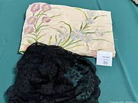 Folded vintage silk embroidered shawl showing colorful floral embroidery with shades of purple, blue, and green, alongside a scrunched black lace scarf on a green surface.