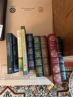 Books arranged standing upright on a cardboard box, showing titles and spines including Pride and Prejudice, The Portrait of a Lady, Leaves of Grass, and others.