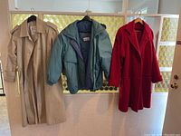 Three ladies coats hanging on hooks: beige trench coat, blue padded jacket, red wool coat