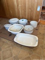 Full view of eight assorted baking dishes, some with lids, placed on floor against wood paneling. Includes Corningware and other brands, with some having blue cornflower pattern and some floral patterns.