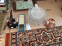 Wide shot showing all items including knives, forks, glass bowls, plates, and decorative jar arranged on cardboard and patterned carpet.