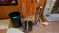 Full view of fireplace tool set on stand, log holder with firewood, black metal bucket and wooden bellows near fireplace.
