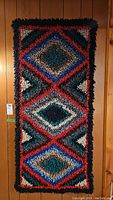 Front view of rectangular handmade geometric shag rug wall art mounted vertically on wood paneling. Shows full pattern of overlapping diamonds in multiple colors.