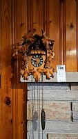Front view of the wooden German cuckoo clock hanging on wood paneling wall above a stone fireplace. Shows the carved leaf and bird details and clock face with Roman numerals.