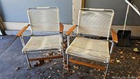 Two vintage lawn chairs with aluminum frames, wood armrests, and white nylon cording sitting on a concrete surface with fallen leaves, showing wear and rust on aluminum.