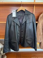 Black leather jacket hanging on a wooden hanger in front of a wooden background. The jacket has a button closure, large collar, and two front pockets.