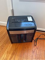 Front and top view of a black Staples desktop paper shredder placed on wooden floor next to a closed white door. Visible control buttons and waste bin.