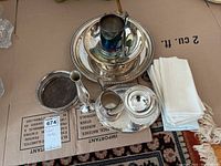 Photo showing multiple silver plated serving pieces including small trays, creamer, covered sugar bowl, and stack of white cloth napkins with tassel ends.