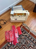 Vintage Singer sewing machine with floral decal, beige hard plastic carrying case, wooden embroidery hoop, assorted spools of thread, and several Aero brand circular knitting needles in red packaging displayed on wooden floor and rug.