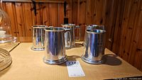 Five metallic pewter and stainless steel beer steins on a table, some with engraved names.