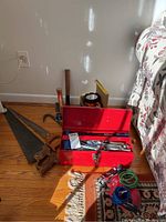 Red metal toolbox with assortment of hand tools including wrenches, pliers, tape measure, hammers, and locks, placed on wooden floor next to a bed.