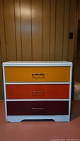 Front view of the vintage three drawer dresser showing the colorful drawers in yellow, orange, and red with metal handles.