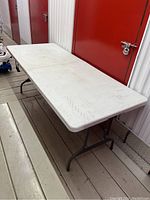 Full-length view of the unfolded table showing white textured plastic top and gray metal legs on wooden floor next to red metal door.