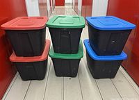 Six black plastic storage bins arranged in two rows of three, each with different colored lids (red, green, blue), standing on wooden floor against red background.