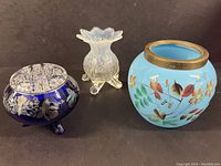 Photo showing three blue glass art pieces: cobalt blue glass flower frog with silver overlay on the left, opalescent ruffled cuspidor in the center, and hand-painted blue vase with brass rim on the right.