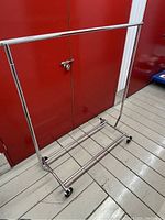 Full view of chrome collapsible garment rack on wheels with horizontal bar and bottom wire shelf against red metal doors.