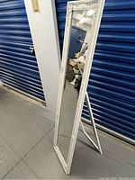 Full view of the full-length standing mirror with carved wooden frame and backing stand in a storage area.