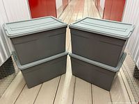 Four grey plastic storage totes with lids stacked in pairs, showing front and side views with wheels visible under bins.