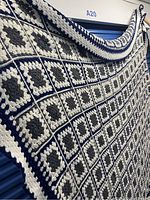 Large crochet bedspread hanging showing many square motifs of gray or black centers, white surrounding, and navy blue outlining with a crocheted border.