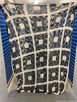 Full view of large crochet bedspread held upright for size reference showing gray, black, and white granny squares connected by white line bands.