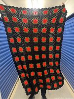 Full view of the black crochet throw blanket with multiple red rose motifs held up showing size and design.