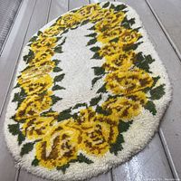 Oval-shaped wool latch hook rug with central cream area surrounded by yellow roses and green leaves, placed on light brown wooden floor.