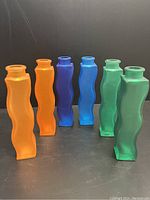 Full set of six vintage IKEA Skamt Vases in orange, blue and green shown standing on black surface with dark background.